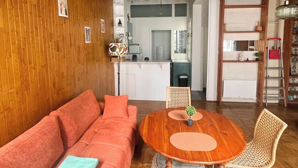 Un accogliente soggiorno con soffitto in legno, un divano rosso, un tavolino rotondo in legno e una poltrona in vimini.