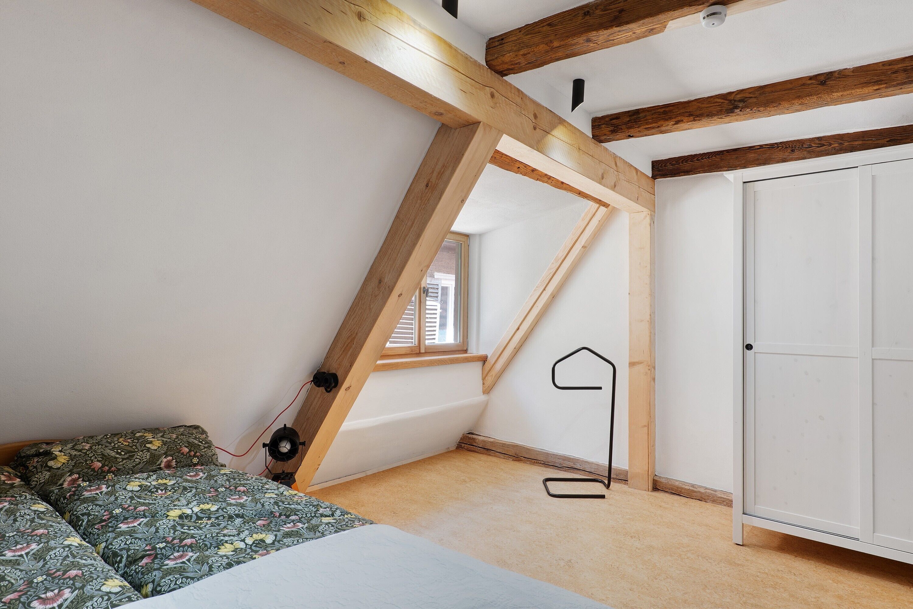A bedroom with a sloped ceiling, a bed with floral bedding, a white wardrobe, and a window with blinds.