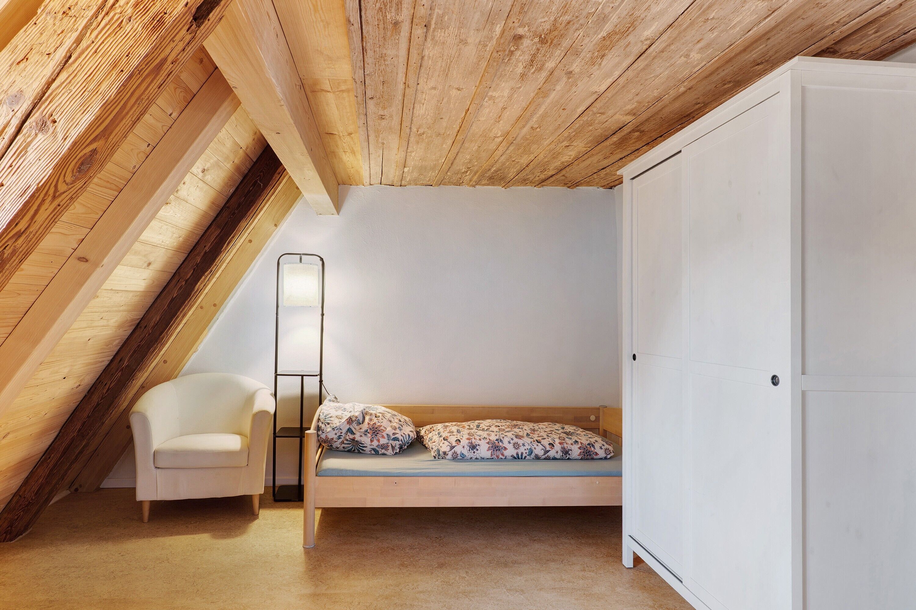 Una camera da letto con soffitto spiovente, un letto con biancheria da letto floreale, una sedia bianca e un armadio.