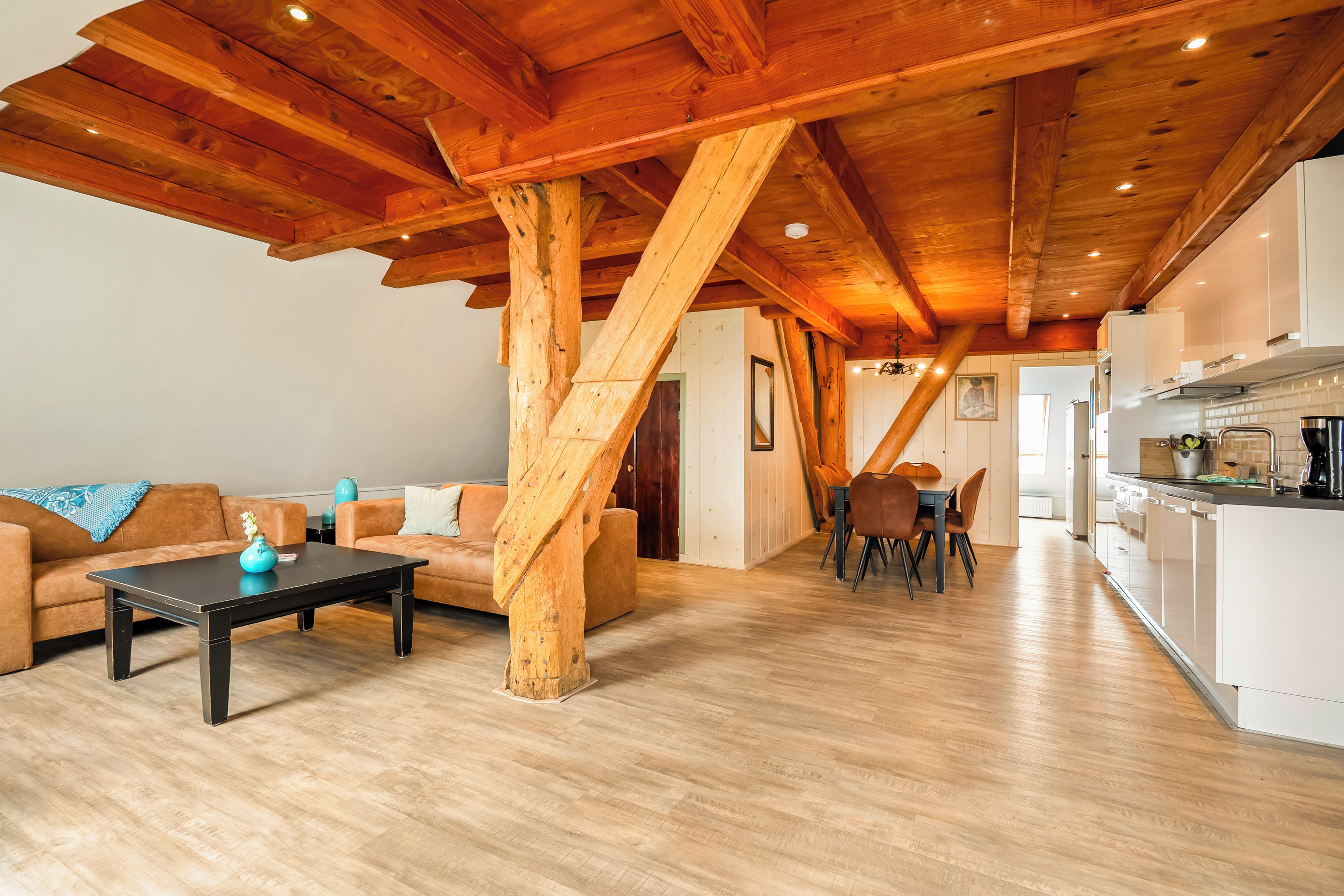Een ruime woonkamer met houten plafond, een zithoek, een salontafel en een keuken in de achtergrond.