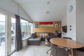 A modern living room with a sectional sofa, a dining table, and a large window with curtains. - A beautiful, detached vacation bungalow in Park Duinland in Sint Maartenszee with a fully fenced-in yard! (Sint Maartensvlotbrug)