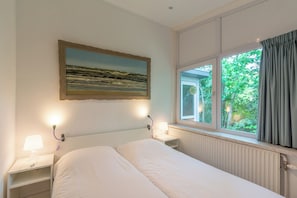 A bedroom with a bed, bedside lamps, a window with curtains, and a painting on the wall. - Beautiful detached holiday bungalow at Duinland holiday park in Sint Maartenszee with a fully enclosed garden! (Sint Maartensvlotbrug)