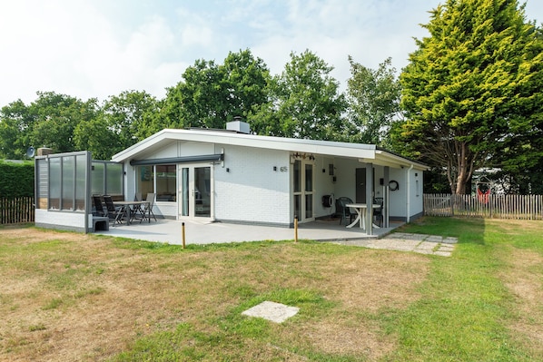 A modern single-story house with a patio area, a covered outdoor seating space, and a visible number '65' on the wall. - A beautiful, detached vacation bungalow in Park Duinland in Sint Maartenszee with a fully fenced-in yard! (Sint Maartensvlotbrug)
