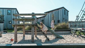 En lekplats med en rutschkana och en klätterställning, omgiven av sand och trähus.
