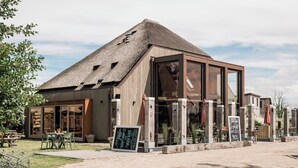En modern byggnad med ett halmtak, stora fönster och sittplatser utomhus.