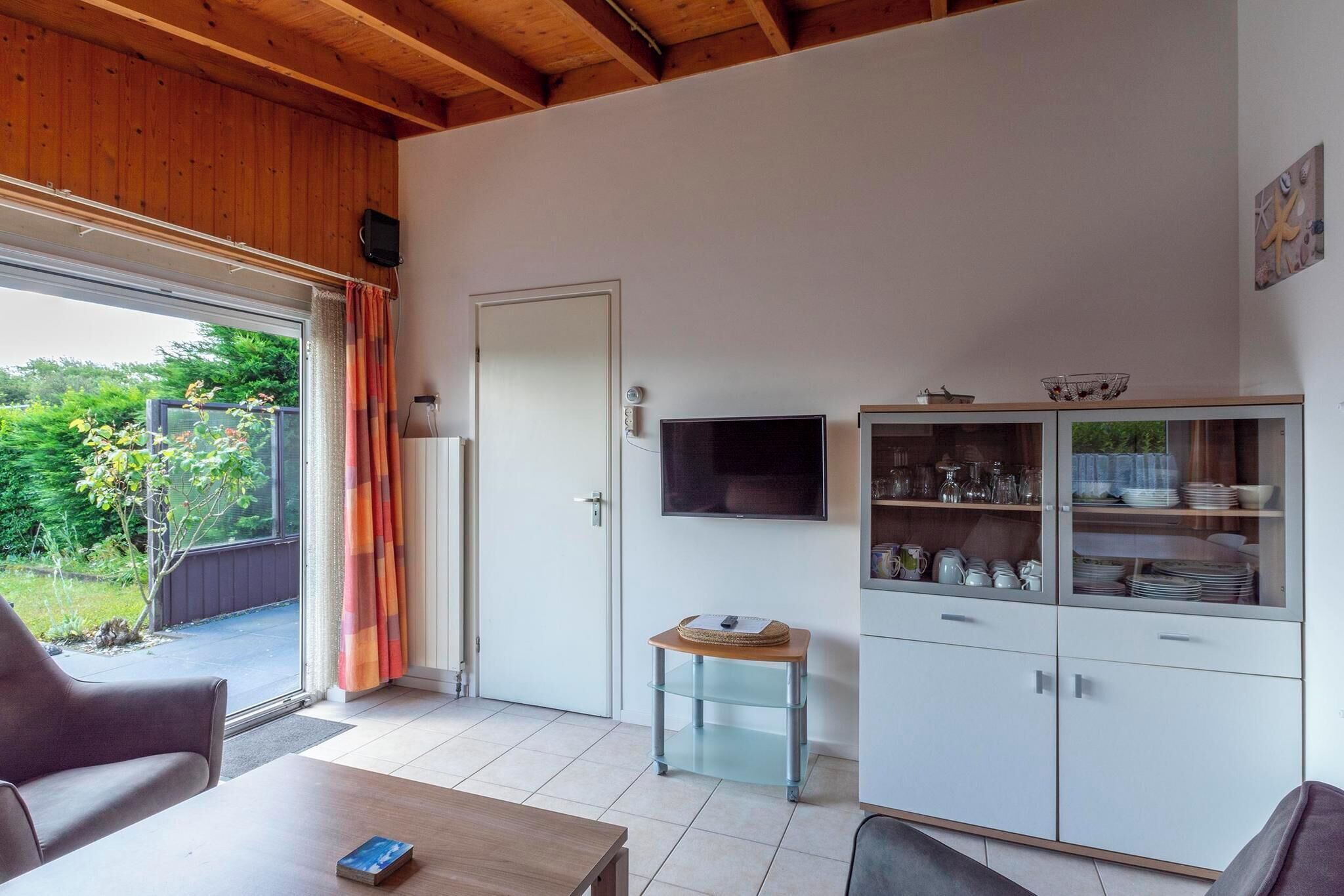 A room with a wooden ceiling, a television mounted on the wall, a cabinet with glass doors, and a table with a book.