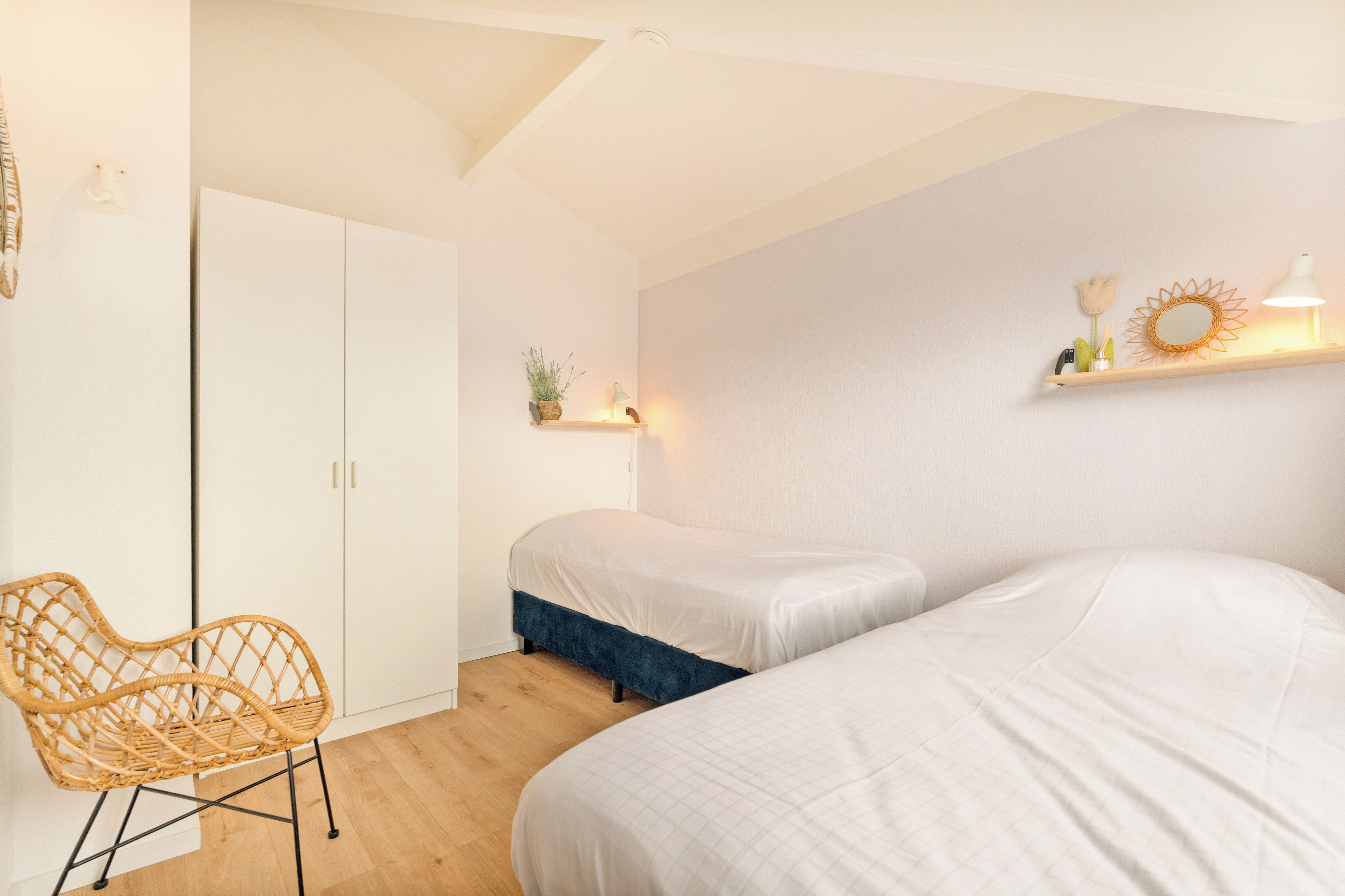 A bedroom with two beds, a chair, a wardrobe, and a shelf with decorative items.