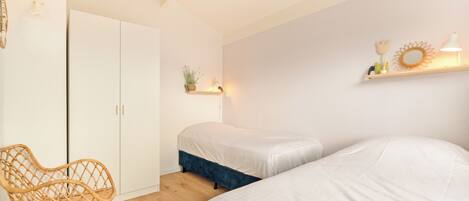 A bedroom with two beds, a chair, a wardrobe, and a shelf with decorative items.