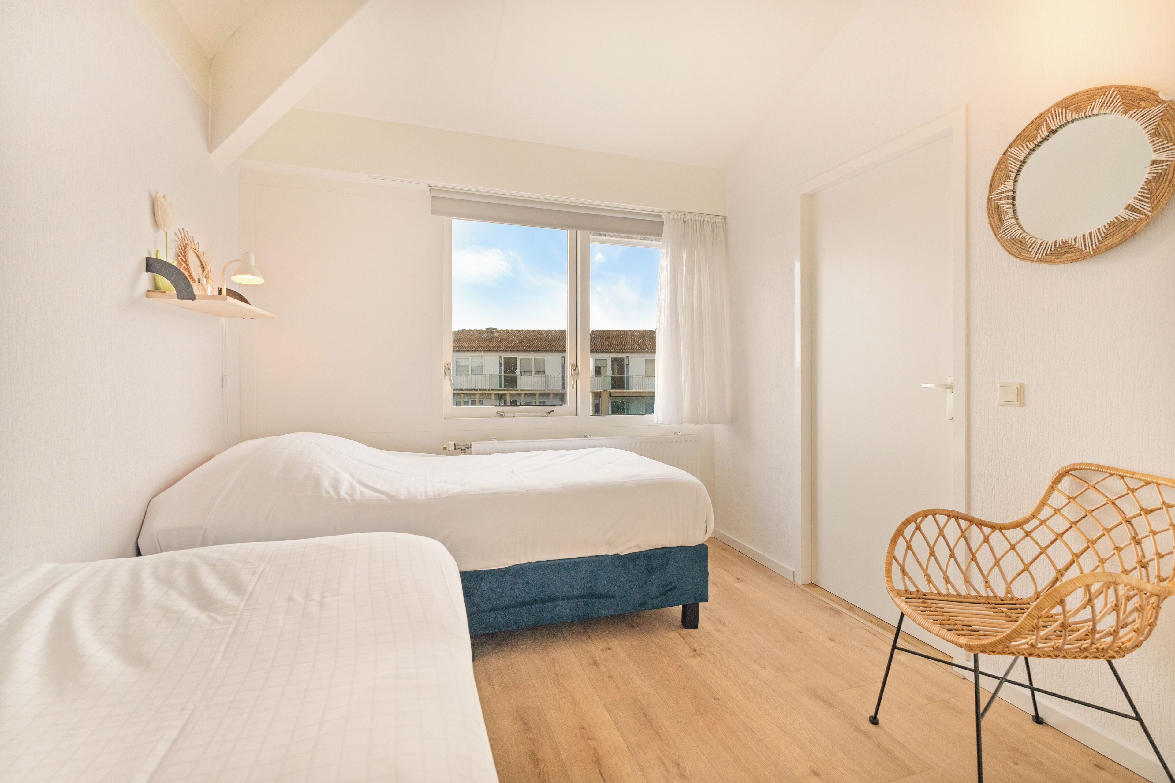 A bedroom with two beds, a window with curtains, a wicker chair, and a wall-mounted mirror.