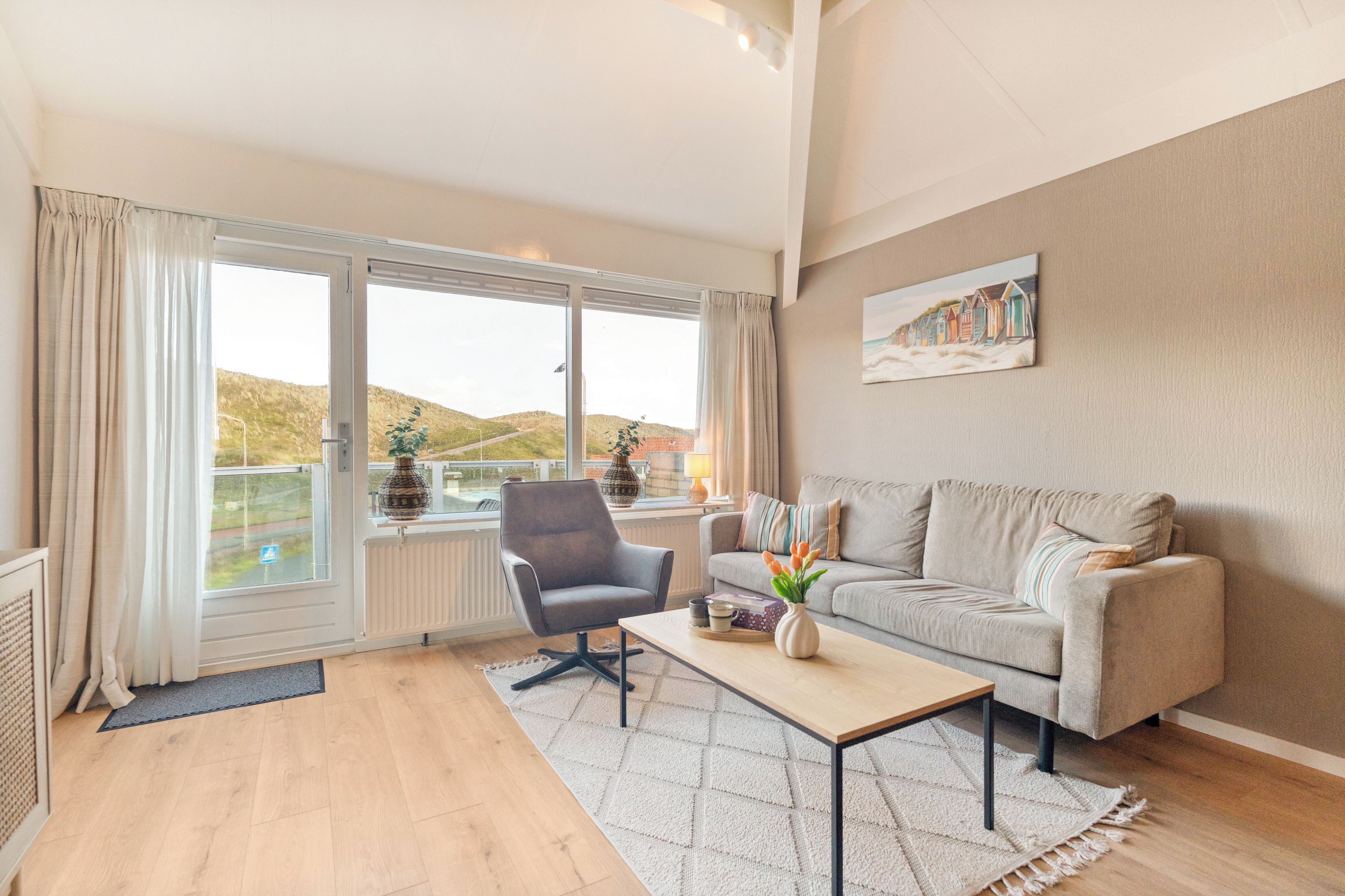 Una sala de estar moderna con un sofá gris, una mesa de centro y una silla. Hay una ventana grande con cortinas, un cuadro en la pared y vistas al exterior.