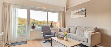 Una sala de estar moderna con un sofá gris, una mesa de centro y una silla. Hay una ventana grande con cortinas, un cuadro en la pared y vistas al exterior.
