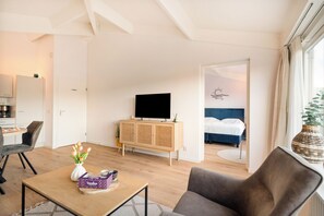 Ein modernes Hotelzimmer mit Holzboden, einem Flachbildfernseher, einem Bett mit blauer Kopfteil, einem Essbereich mit Tisch und Stühlen und einem großen Fenster mit Vorhängen.