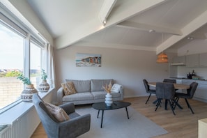 Ein modernes Wohnzimmer mit Sofa, Couchtisch und Esstisch. An der Wand hängt ein Gemälde und es gibt ein Fenster mit Vorhängen.