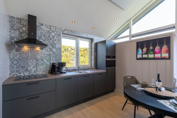 A modern kitchen with a large window, a built-in stove, and a dining area with a table set for two. - Luxury Wellness Villa for 2 People (Julianadorp)