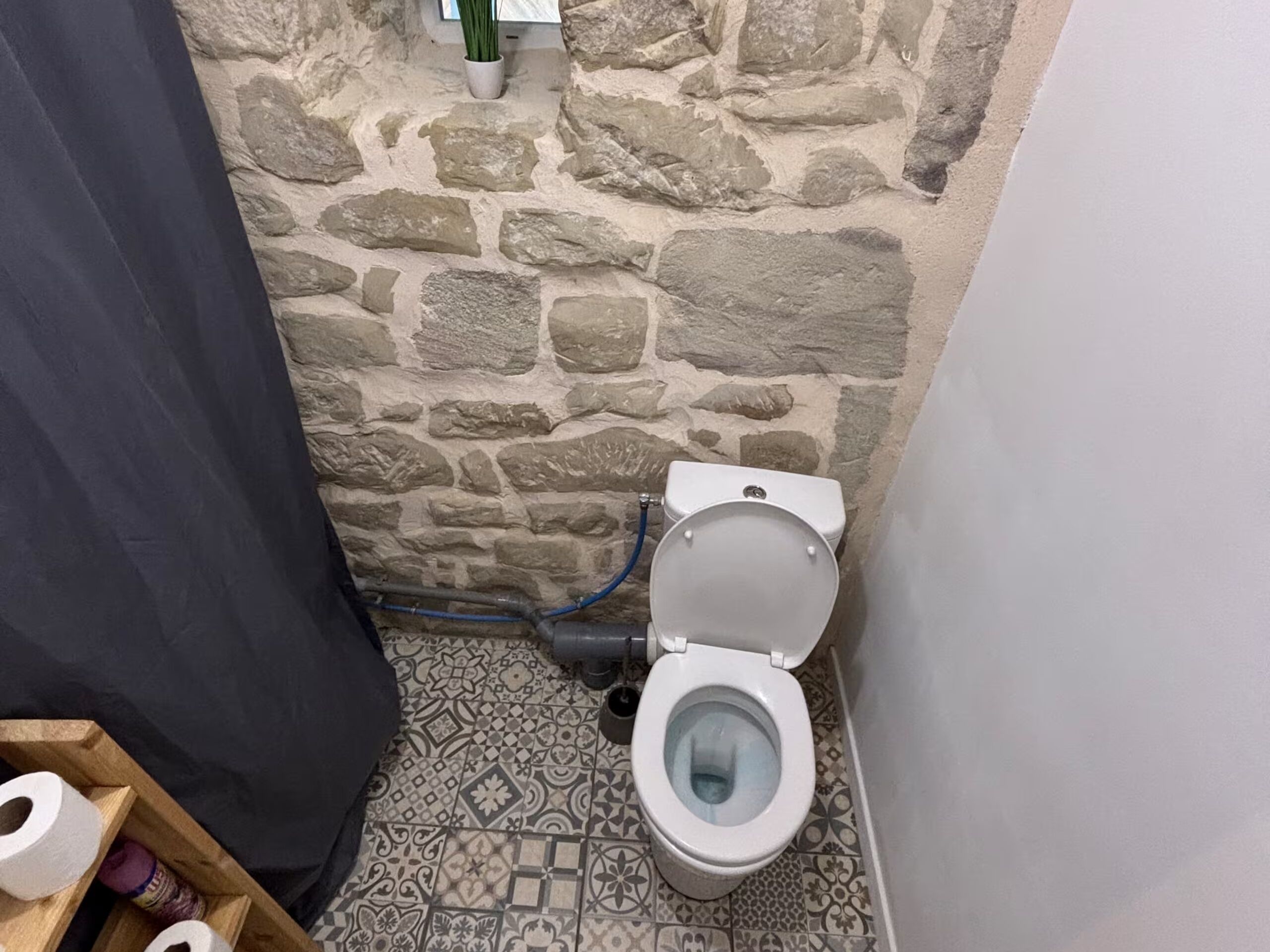 A bathroom with a stone wall, a toilet, patterned floor tiles, and a wooden shelf with toilet paper.