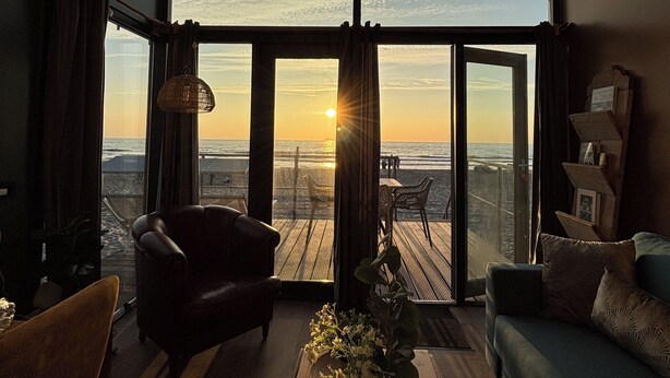 Een kamer met een grote glazen deur die leidt naar een balkon met uitzicht op de ondergaande zon boven de oceaan.