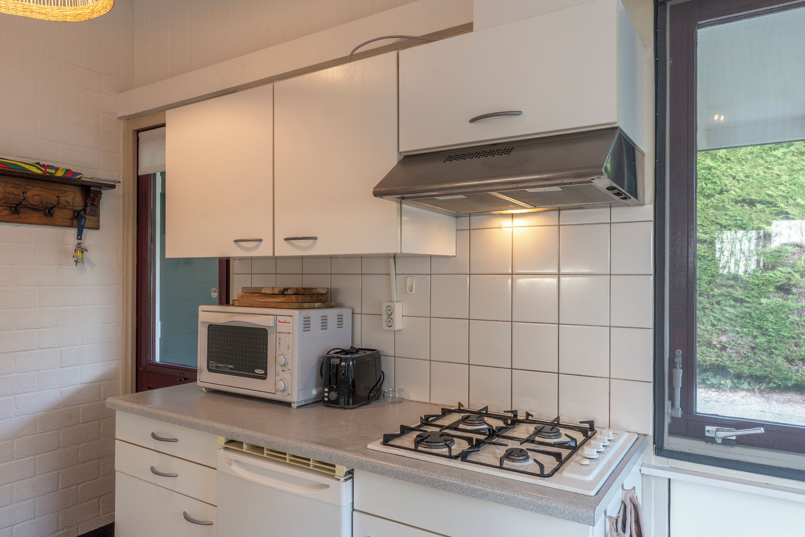 Eine Küche mit weißen Schränken, einer Mikrowelle, einem Toaster, einem Gasherd und einem Fenster mit Blick auf Grünflächen.