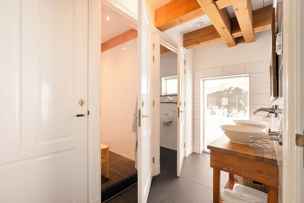 Une salle de bain carrelée de carreaux blancs, avec un meuble-lavabo en bois, un évier et une fenêtre équipée de stores.