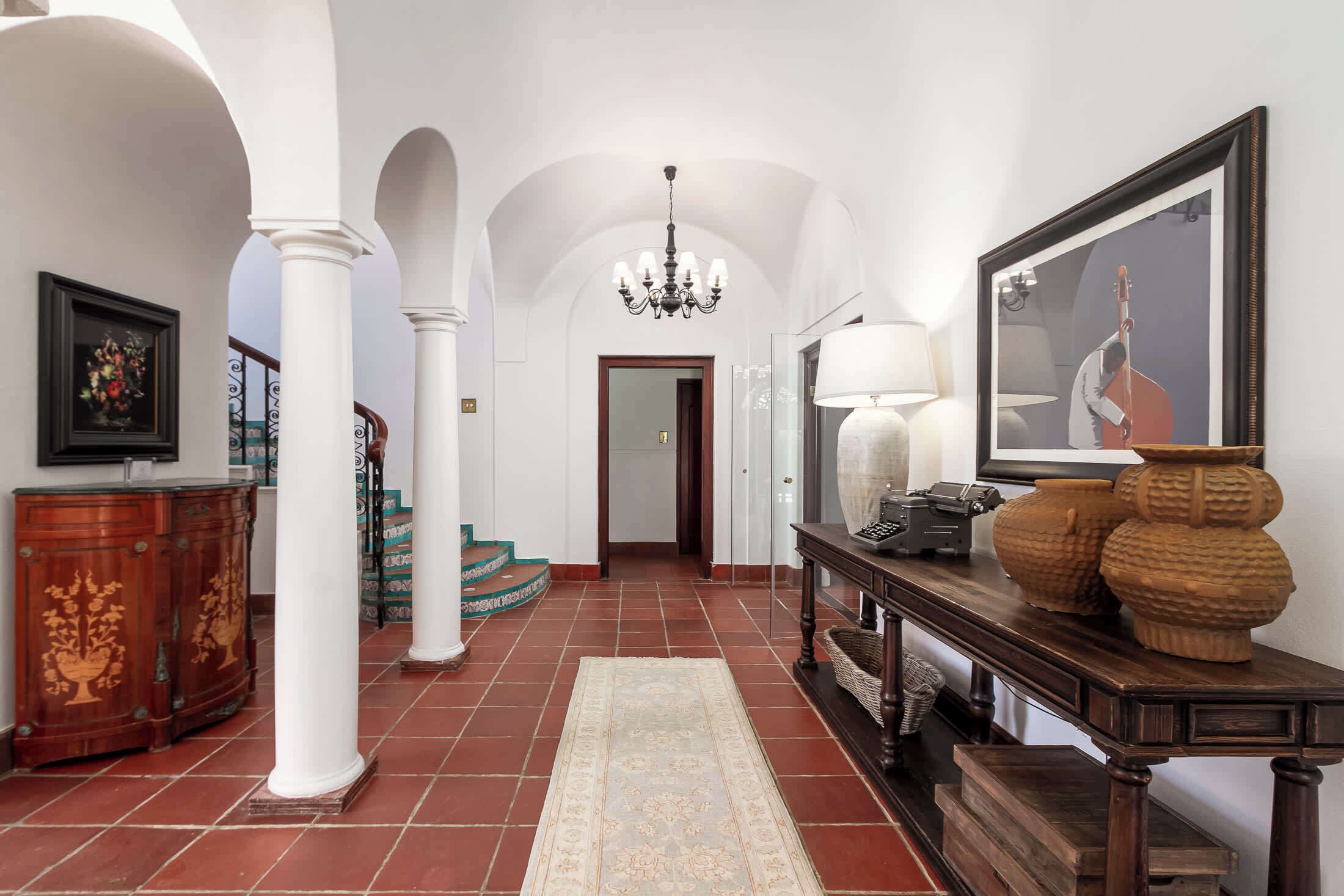 Un vaste hall d’entrée avec des colonnes blanches, un lustre, une console en bois agrémentée d’objets décoratifs et un tableau encadré au mur.