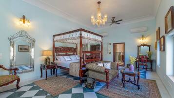 A spacious bedroom with a four-poster bed, a sitting area with a sofa and armchairs, a chandelier, and a checkered floor.