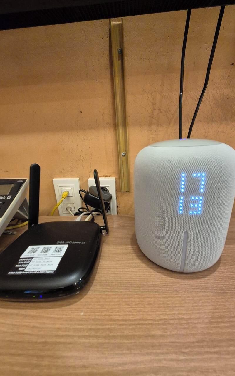 A router and a smart speaker on a wooden surface with a wall-mounted electrical outlet in the background.