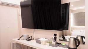 Quarto de hotel com televisão de tela plana, mesa de cabeceira com chaleira, cafeteira e espelho refletindo o ambiente.