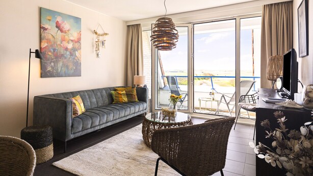 A modern living room with a sofa, a round table, wicker chairs, a painting, and a view through a glass door. - A modernly furnished vacation rental with a view of the dunes of Groote Keeten. (Callantsoog)