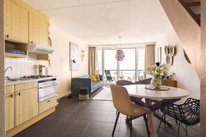 Eine moderne Küche mit Holzschränken, eine Essecke mit einem runden Tisch und ein Blick durch eine Glastür auf einen Außenbereich mit Sitzgelegenheiten.