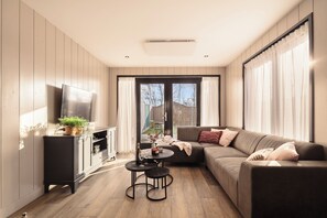A modern living room with a sofa, a TV, a small table with a bottle and glasses, and a cabinet with plants. - Zand Vacation Home – Luxury, Peace, and Space on the Coast (Julianadorp)