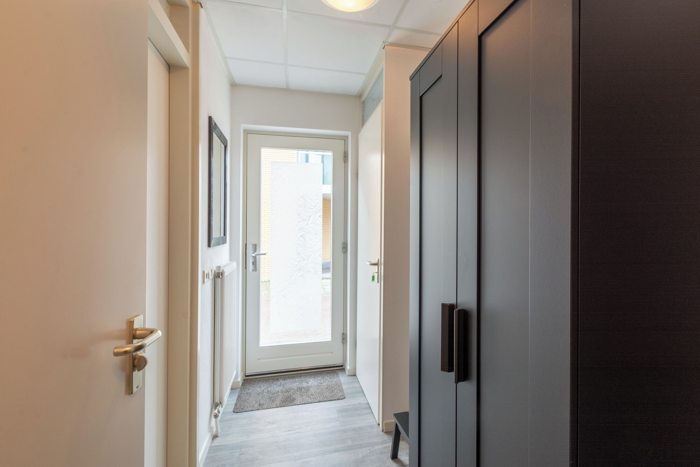 Een gang met een deur die leidt naar een lichte kamer, een donkere deur en een vloer in een lichte tint.