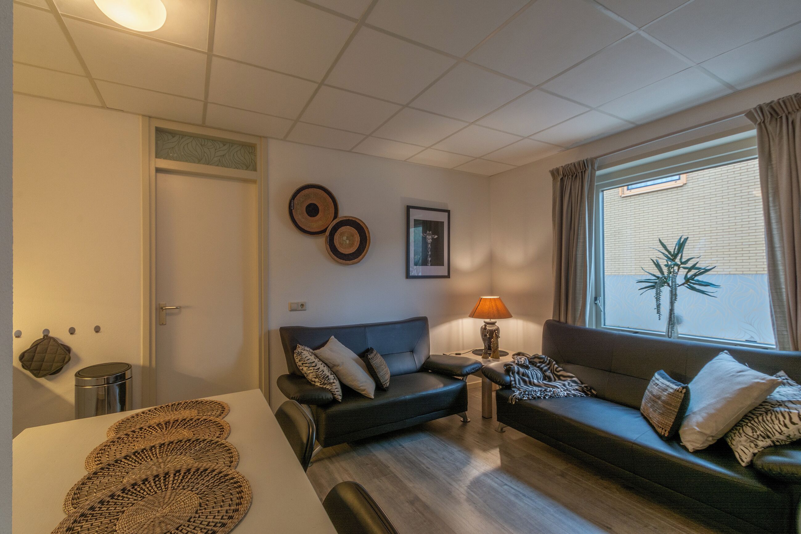 Een woonkamer met een bank, een tafeltje met een lamp, een prullenbak en een raam met jaloezieën.
