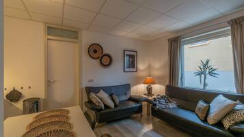 Een woonkamer met een bank, een tafeltje met een lamp, een prullenbak en een raam met jaloezieën.
