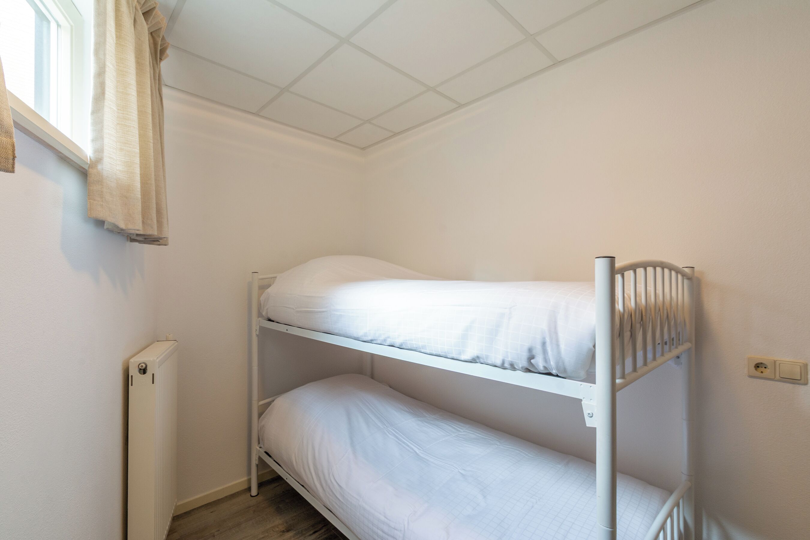 A bunk bed room with a radiator, a window with a curtain, and a switch on the wall.