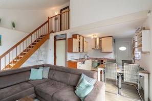 Eine moderne Wohnung mit einer Holztreppe, einem braunen Sofa, einem Essbereich mit Korbstühlen und einer Küche mit weißen Schränken.