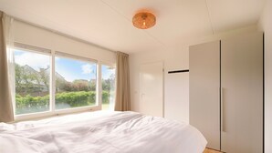 A bedroom with a large bed, a wardrobe, and a window with a view of greenery. - A beautiful, modernly furnished vacation rental for 4 people, located right across from the beach entrance in Callantsoog! (Callantsoog)