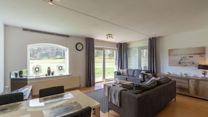 Ein modernes Wohnzimmer mit Sofa, Esstisch und Blick auf einen Garten durch ein großes Fenster.