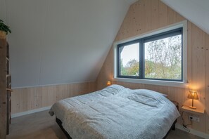 A bedroom with a sloped ceiling, a large window, a bed, a bedside lamp, and a wooden wall. - Modern, newly furnished detached villa for 8 guests, with a lovely large garden (Groote Keeten)