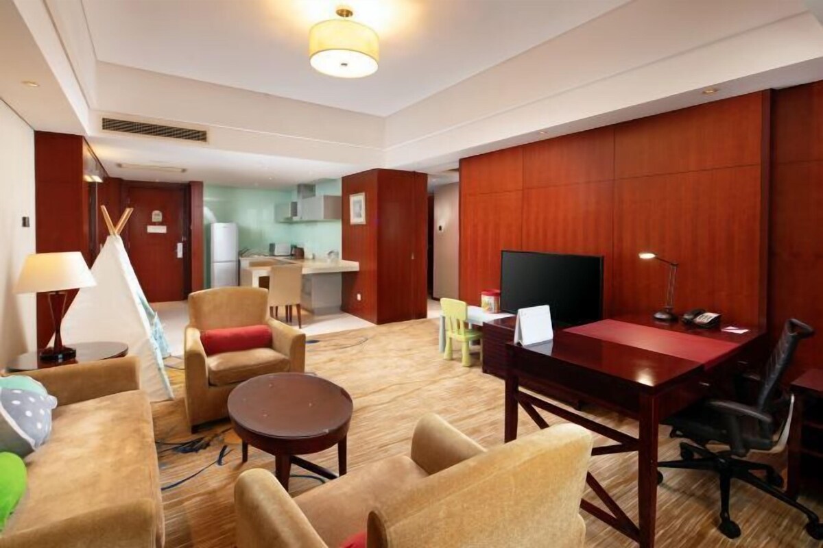 A hotel room with a dining table, chairs, a TV, and a kitchenette in the background.