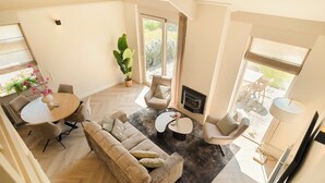 A spacious living area with a sofa, coffee table, and dining table, a fireplace, and large windows offering a view of the outdoors. - This beautiful detached vacation home in Julianadorp aan Zee is fully equipped with all modern conveniences. (Julianadorp)