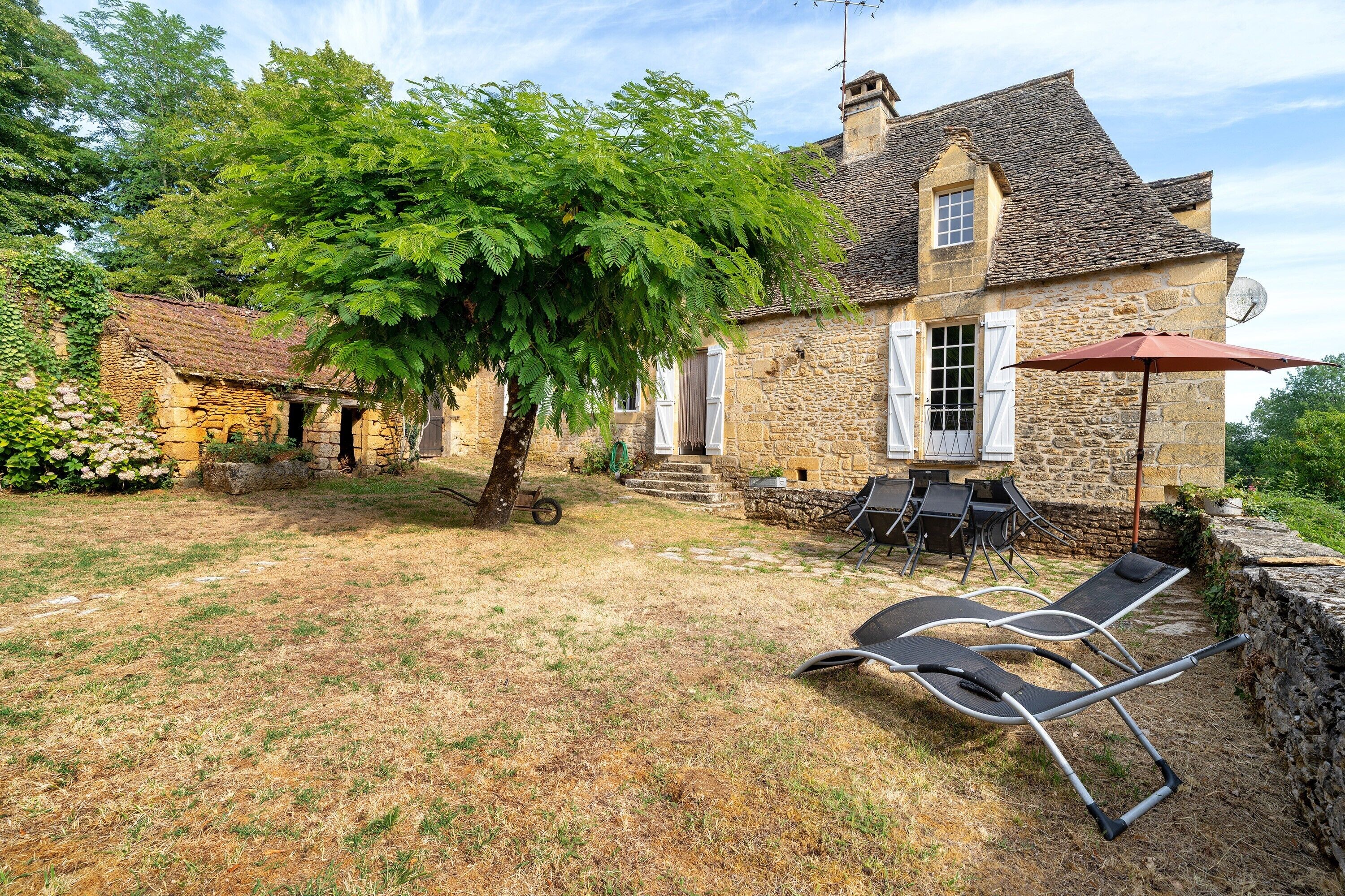 A stone house with a tree in the yard, outdoor furniture, and a satellite dish.
