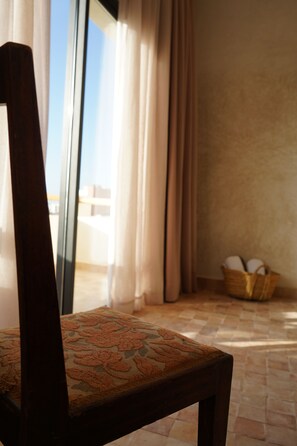 A wooden chair with a patterned cushion, a window with curtains, and a wicker basket on the floor.