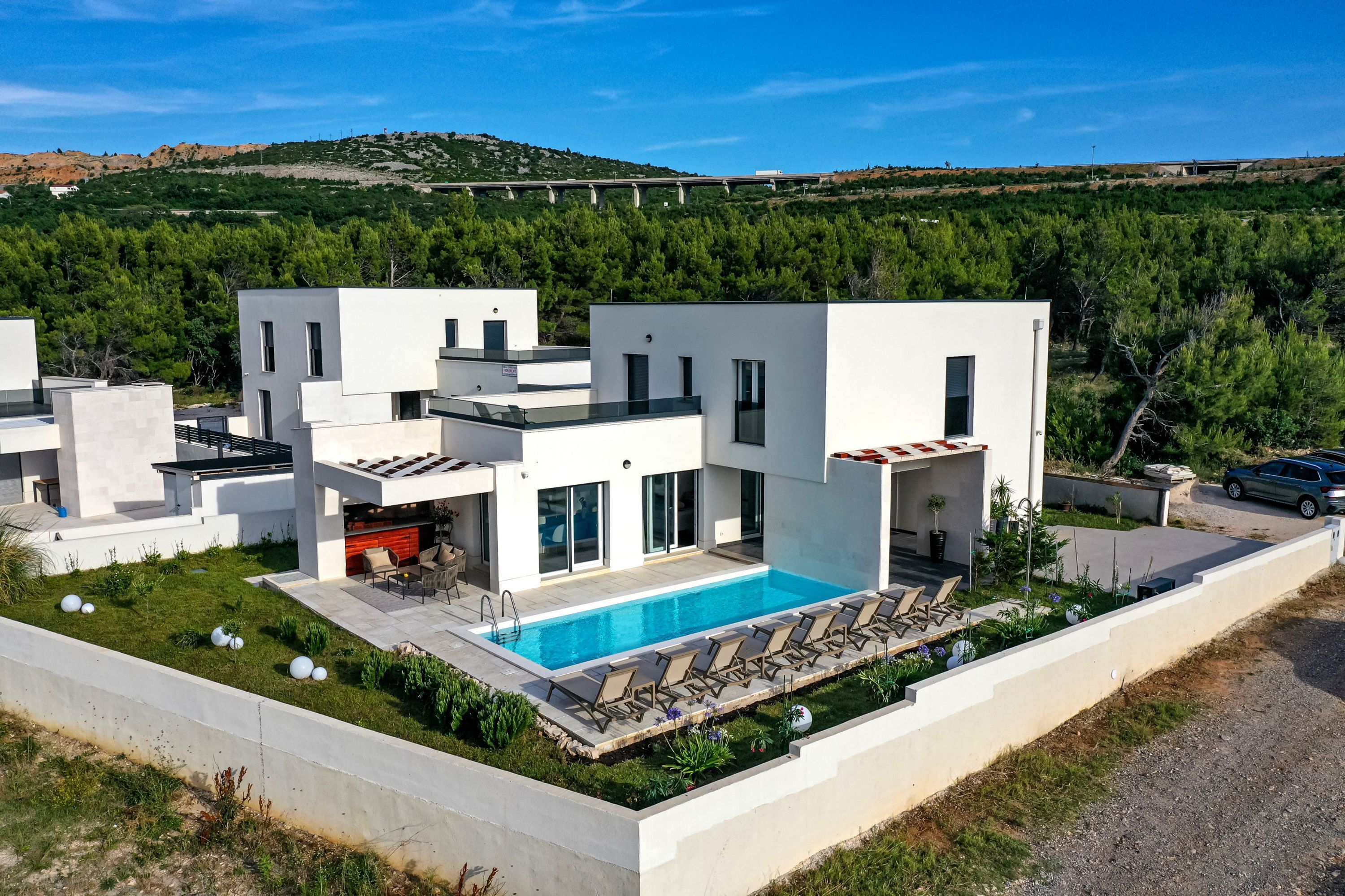 A modern villa with a swimming pool, multiple balconies, and a view of a bridge and forested hills.
