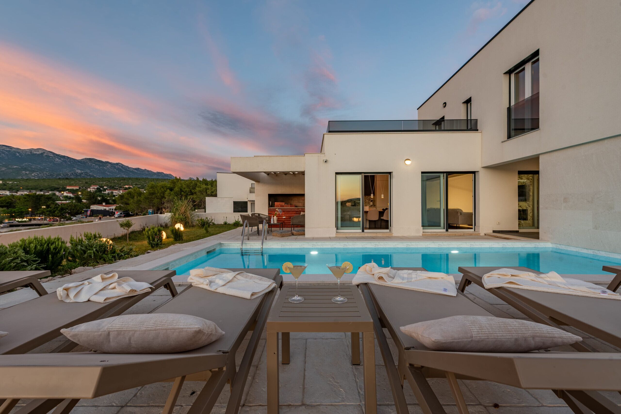A modern outdoor pool area with lounge chairs and a table, a building with large windows, and a view of mountains in the background.