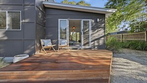 Une maison moderne dotée d’une terrasse en bois, de deux fauteuils en osier et d’une porte en verre donnant sur un espace intérieur.