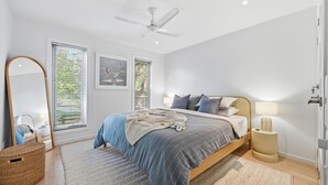 Une chambre à coucher comprenant un lit, un ventilateur de plafond, une lampe de chevet, un miroir et une fenêtre avec des stores.