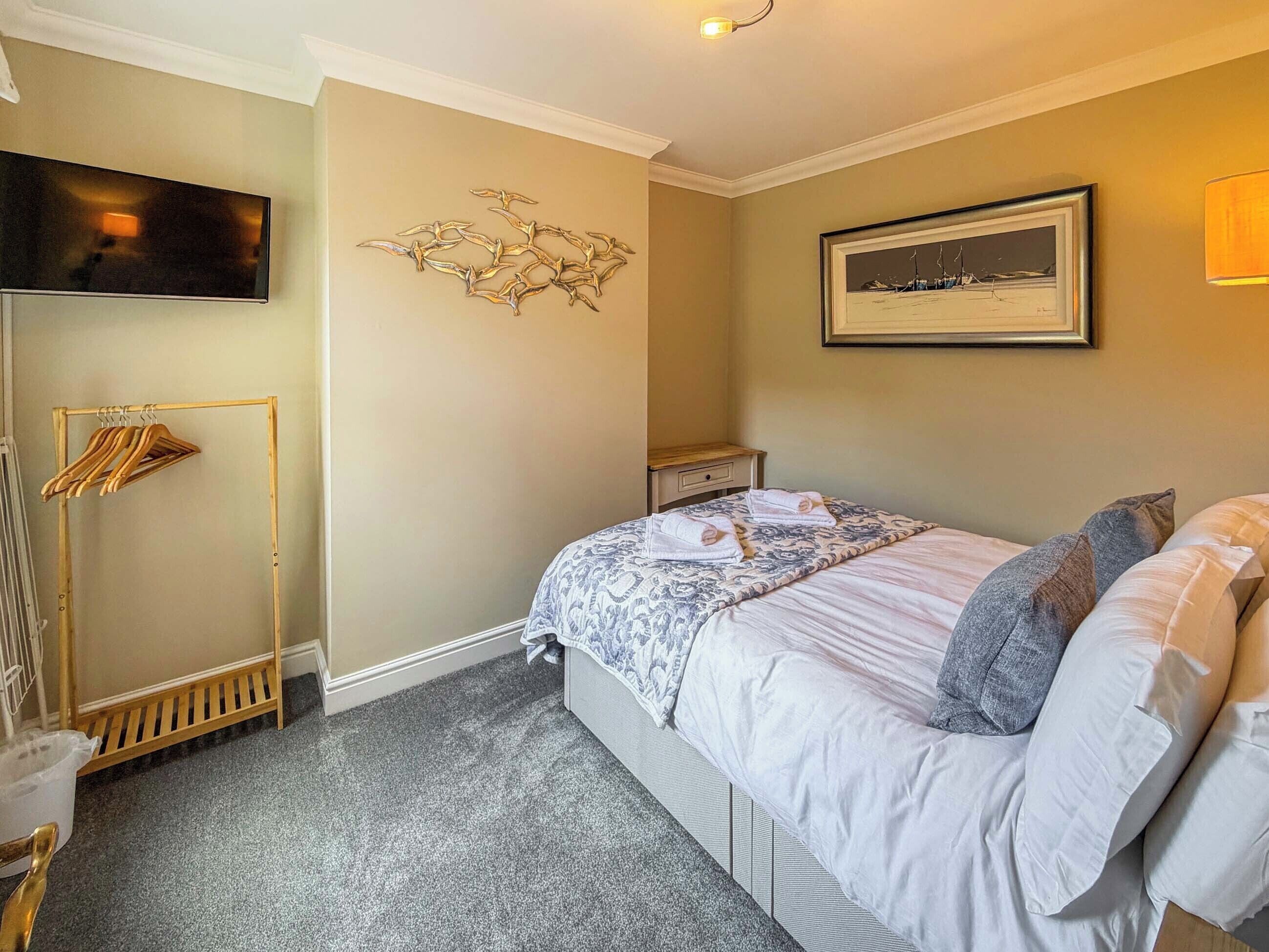 Een slaapkamer met een bed, een houten kledingrek, een ingelijst schilderij en een televisie aan de muur.
