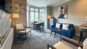 A living room with a sofa, chairs, a table, a radiator, a clock, and a painting.