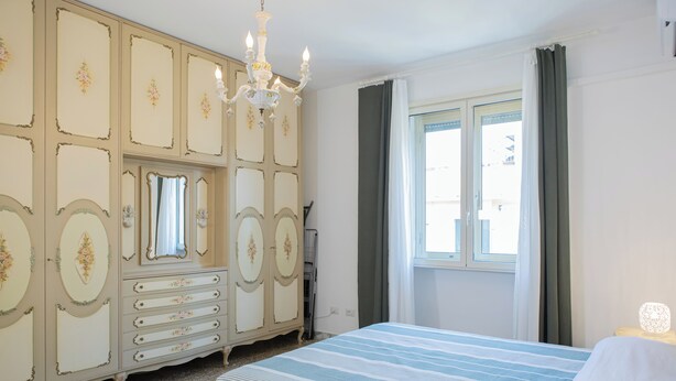 Un dormitorio con cama, cómoda, espejo, candelabro, ventana y cortinas.