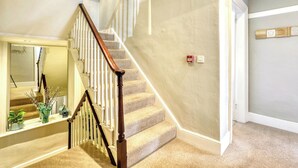 Una escalera con alfombra, barandilla de madera y balaustres blancos, que conduce a una habitación con un espejo y una planta pequeña en el alféizar de la ventana.