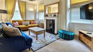 Una sala de estar con sofá, reposapiés, mesa de centro, chimenea, televisor y cortinas.